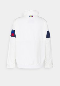 Chaqueta blanca con cuello alto, detalles en azul marino en las mangas y parches de colores en la parte superior de la espalda. Fabricada con una tela suave.