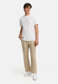 Camiseta de algodón blanca con un pequeño logo gráfico en el pecho, combinada con pantalones beige claros y zapatillas blancas, creando un look casual.