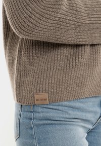 Bruine gebreide trui met ribbelstructuur, met een logo-opdruk aan de onderkant, in combinatie met lichtblauwe spijkerbroek.