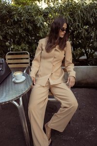 Costume beige ajusté avec une veste cintrée et un pantalon à jambes larges, associé à des lunettes de soleil noires, une table en argent et une tasse de café.