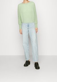 Ljusgrön, texturerad stickad tröja med rund hals, kombinerad med ljusblå raka jeans och svarta plattformsträskor, stående på en neutral bakgrund.