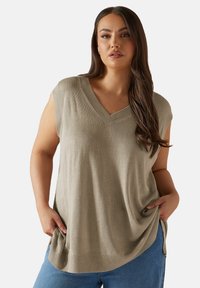 Ljusgrön ärmlös topp med v-ringning, tillverkad av strukturerad tyg, med lös passform och sidofötter, ihop med blå jeans.