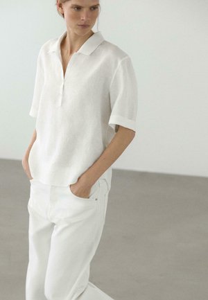 Donna con indosso una camicia bianca a maniche corte e collo, ampia, e pantaloni bianchi, in piedi con le mani in tasca contro uno sfondo grigio uniforme.