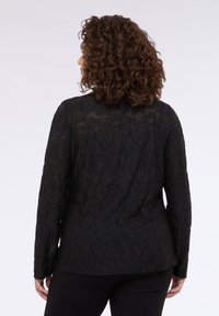Langärmliges schwarzes Oberteil mit strukturiertem Blumenmuster, transparenter Überlagerung und rundem Ausschnitt, kombiniert mit schwarzen Hosen.