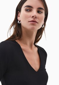 Donna con lunghi capelli castano che indossa orecchini a cerchio argentati intrecciati e una maglietta nera con scollatura a V, che guarda di lato su uno sfondo bianco.