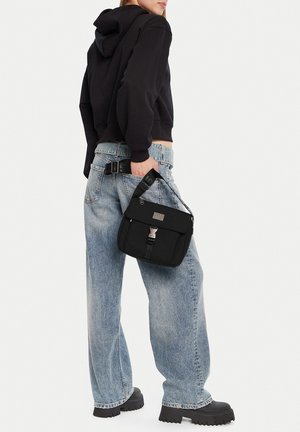 Person trägt schwarzen Hoodie und weite blaue Jeans, hält eine schwarze Schultertasche mit Schnappverschluss, steht vor weißem Hintergrund.