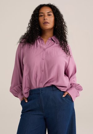 Vrouw met krullend haar die een losse roze blouse met knopen en blauwe hoog getailleerde broek draagt, staand met handen in de zakken tegen een effen achtergrond.