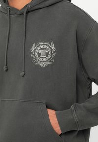 Sötétszürke kapucnis pulóver elülső zsebével, húzózsinórral, és egy kis "Tommy Jeans New York" logóval a mellkason. Kéz a zsebben.