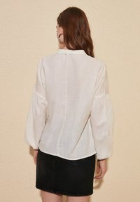 Lette, elfenbensfarvet bluse med puffede ærmer og rund kant. Teksturstof med en subtil glans. Sættes sammen med en sort denim-nederdel.