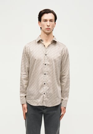 Lühikeste tumedate juustega mees kannab beeži mustrilist nööbitavat särki ja tumedat halli pükse, seistes ühevärvilise heleda taustaga.