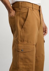 Ruskeat cargo-housut, joissa on useita sivutaskuja, valmistettu kestävästä kankaasta, jossa on ommeltuja yksityiskohtia ja metallinen napsulukko.