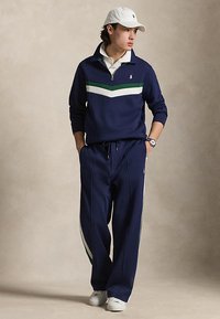 Jonge man in marineblauw trainingspak met witte en groene strepen, witte sneakers, wit poloshirt, witte pet en horloge, staand met handen in de zakken.