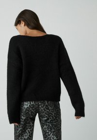 Zwarte, oversized trui met een brede halslijn en lange mouwen, gemaakt van een textuurgebreide stof. Gecombineerd met zwarte broek met luipaardprint.
