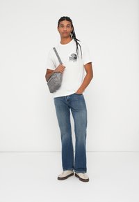 Mladý muž s dlouhými pletenými vlasy, oblečený v bílém tričku s potiskem, modrých džínách, béžových teniskách a nese šedou crossbody tašku.