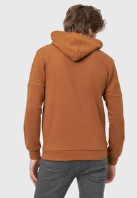 Sweat à capuche marron avec des panneaux d'épaules matelassés texturés, des poignets et un ourlet côtelés. Tissu lisse avec une coupe décontractée, montré de dos.