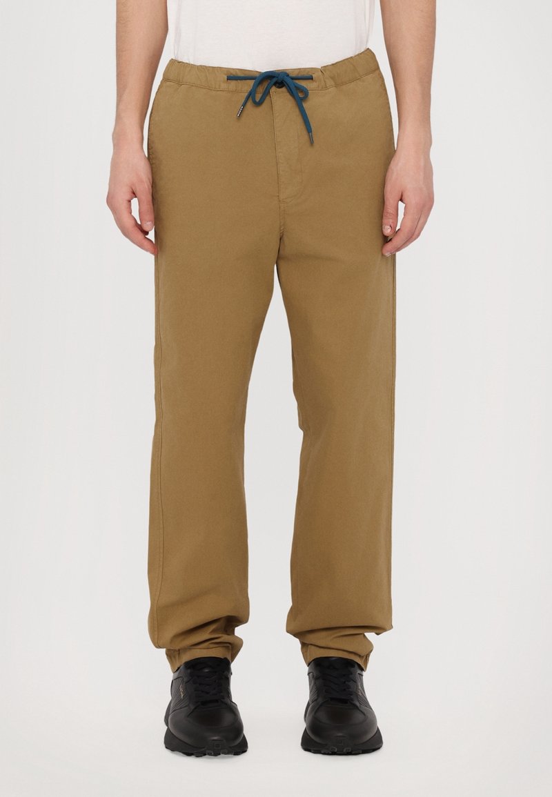 Homme portant un pantalon beige à cordon de serrage avec taille élastique et des baskets noires, debout contre un fond neutre.