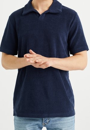Homme portant un polo bleu foncé à manches courtes et un jean bleu clair, les mains jointes devant à hauteur de la taille.