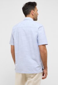 Koszula zapinana na guziki, z krótkim rękawem, w jasnoniebieskim kolorze, wykonana z teksturowanej tkaniny; ma prosty kołnierzyk i boczne rozcięcia dla swobody ruchów.