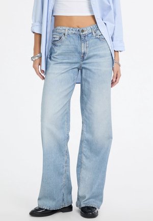 Femme portant un jean bleu clair à jambes larges, des chaussures noires, un top blanc court et une chemise bleu clair boutonnée aux manches retroussées.
