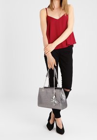 Grijze handtas met een gestructureerde afwerking, voorzien van zilveren kettinghandvatten en een decoratieve hanger, vastgehouden door een persoon in een rode top en zwarte broek.