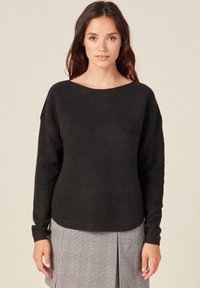 Schwarzer Strickpullover mit weitem Ausschnitt und langen Ärmeln, versehen mit einem strukturierten Muster, kombiniert mit einem karierten Rock.