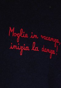 Frase italiana ricamata in rosso "Moglie in vacanza, inizia la danza!" su tessuto blu scuro.