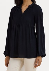 Kvinna i en långärmad svart blus med veck och knapppdetaljer vid halsringningen, i kombination med ljusbeiga byxor.