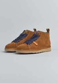 Un paio di stivali alla caviglia in suede marrone con lacci blu scuro, suole beige e fodera interna in maglia, esposti su uno sfondo grigio.