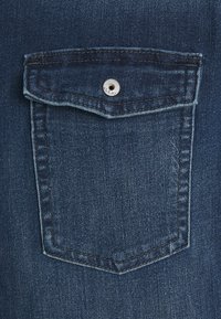 Denim pocket with a flap, metallic snap closure, dark blue color, and visible stitching details; fabric has a smooth, textured appearance.