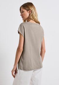 Vaaleanbeige lyhythihainen t-paita, jossa on rento istuvuus, sileä tekstuuri, pyöreä kaula-aukko ja hieman kaareva helma, yhdistettynä valkoisiin housuihin.