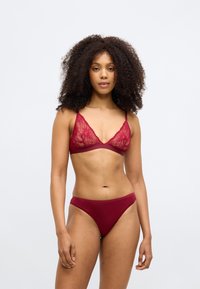 Crvena čipkasta braletica s tankim naramenicama i dubokim dekolteom, uparenim s crvenim bikini gaćicama, koje imaju glatku tkaninu i minimalan dizajn.