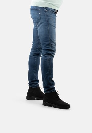 Lässige blaue Jeans mit Slim Fit, die sichtbare Nähte und eine Reißverschlusstasche aufweisen. Getragen mit schwarzen Wildleder-Stiefeletten.