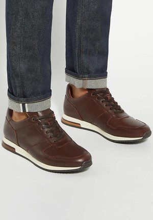 Sneakers in pelle marrone con suole bianche indossate con jeans blu scuro risvoltati su sfondo bianco.