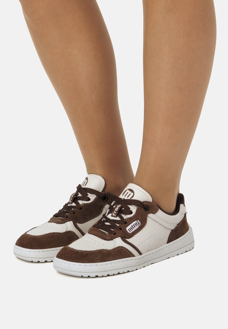 Person, die weiße und braune Sneaker mit Schnürsenkeln trägt, steht mit eng zusammenstehenden Füßen vor einem einfarbigen Hintergrund.