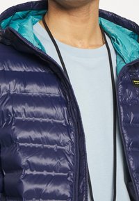 Uomo che indossa una giacca a sacco blu navy con zip e fodera teal sopra una camicia azzurra chiara, con una collana nera in corda visibile attorno al collo.