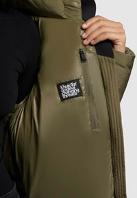 Person wearing black top holding open olive green padded jacket with inner zip pocket and "Khujo" brand label visible.