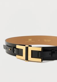 Μαύρη δερμάτινη ζώνη με ορθογώνιο αγκράφα σε χρυσό τόνο και διακοσμητικά στοιχεία. Διαθέτει λεία υφή και αντίθετη εσωτερική επένδυση σε καφέ χρώμα.