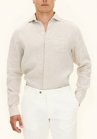 Camicia a maniche lunghe beige chiaro in tessuto testurizzato, con chiusura a bottoni frontale e taschino sul petto, abbinata a pantaloni bianchi.