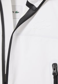 Giacca bianca realizzata in un tessuto liscio e lucido, caratterizzata da una cerniera nera e dettagli in nero, logo impresso e design minimalista.