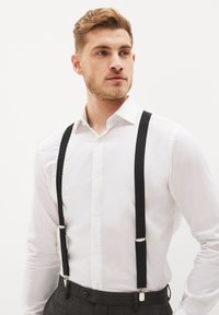 Tirantes de tela negra con clips plateados llevados sobre una camisa blanca de botones, combinados con pantalones de cuadros grises. Diseño sencillo y funcional.