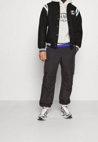 Zwarte varsity jas met witte en groene strepen, grijze hoodie met tekst, donkere cargobroek en zilveren sportschoenen. Casual gelaagd outfit.