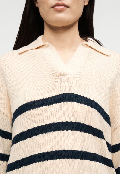Γυναίκα που φοράει μπεζ πλεκτό πουλόβερ με γιακά και τρεις μαύρες οριζόντιες ρίγες στο στήθος και στα μανίκια.