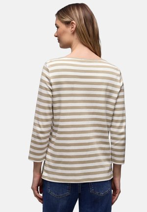 Frau mit hellbraunen Haaren, die ein langärmeliges Hemd mit beigen und weißen Streifen und blaue Jeans trägt, von hinten vor einem schlichten Hintergrund gezeigt.