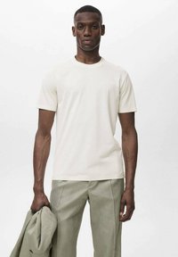 T-shirt a maniche corte, collo rotondo, in tessuto bianco sporco, con texture liscia, abbinato a pantaloni verde chiaro, modello che tiene in mano una giacca coordinata.