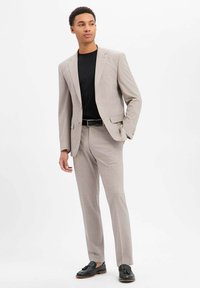 Hellgrauer Anzug mit einem Zweiknopf-Blazer und flachgeschnittenen Hosen. Getragen über einem schwarzen T-Shirt, kombiniert mit schwarzen Loafers. Glatte Textur.