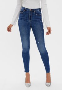 Högmidjade blå denimjeans med en figurnära design, lätt slitage och två framfickor, kombinerat med silverfärgade högklackade skor.