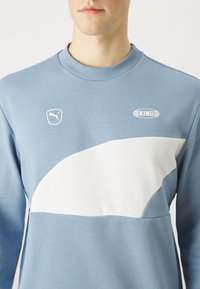 Sudadera azul con panel curvado blanco, que presenta un pequeño logo de Puma y el emblema "KING". Diseño de cuello redondo, textura de tela suave.