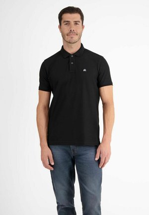 Mann mit kurzen dunklen Haaren, der ein schwarzes Polohemd mit kleinem Logo und blaue Jeans trägt, vor einem einfarbigen weißen Hintergrund stehend.