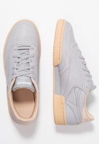 Graue Leder-Sneaker mit einer hellbraunen Gummisohle. Merkmale sind eine perforierte Zehenpartie, eine Schnürung und ein Logo auf der Zunge.