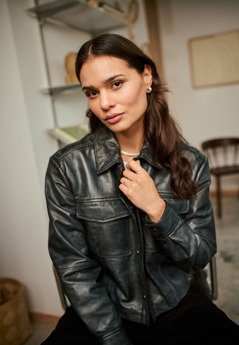 Zwarte leren jas met een knoopsluiting aan de voorkant, twee borstzakken en een spitse kraag. Model met lang haar poseert binnenshuis.